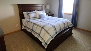 A bedroom with a dark wood bed, a plaid bedspread, a nightstand with a lamp, and a window with blue curtains. - Convenient to Green Bay's beltline where you can get anywhere in 15 minutes. (Green Bay)