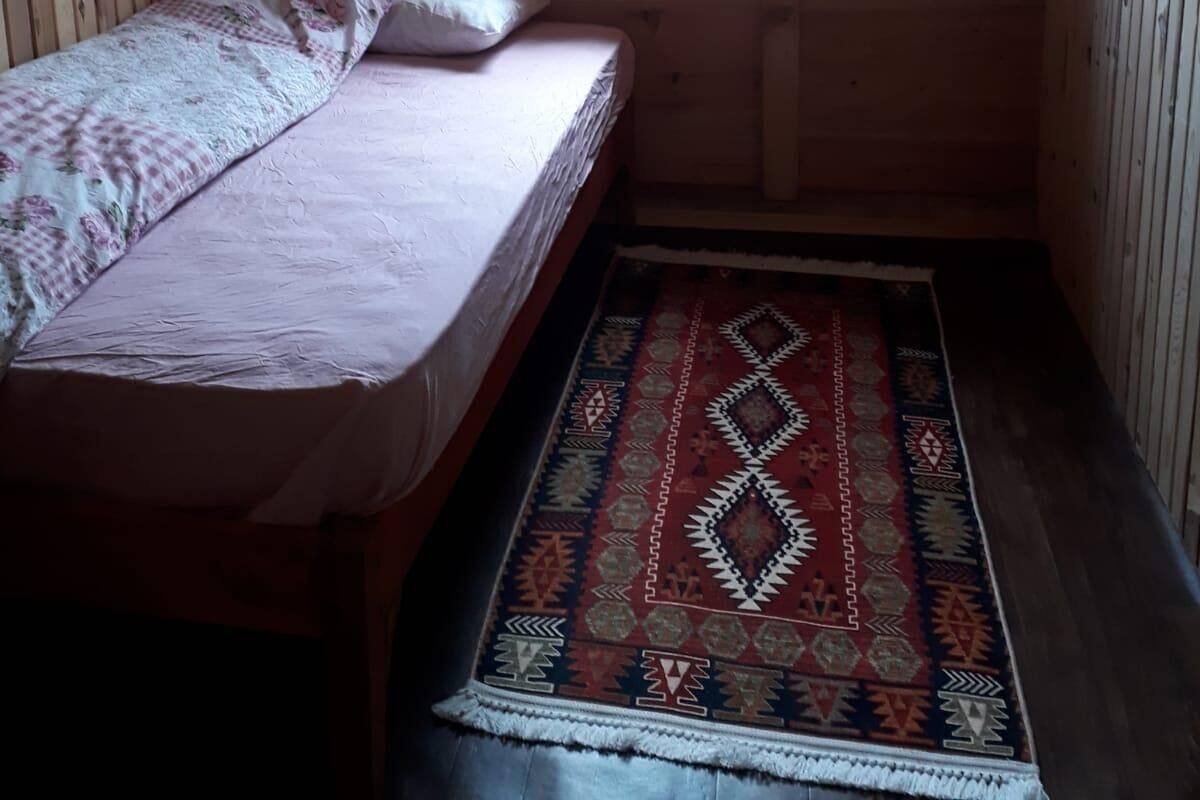 Quarto pequeno com cama de solteiro, teto inclinado e janela. Um tapete estampado está no chão.