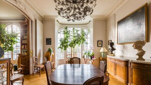 Een eetruimte met een grote houten tafel, stoelen, een kroonluchter, een boekenkast en een groot raam met uitzicht op de straat.