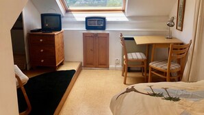 Ein kleiner, gemütlicher Raum mit schrägen Wänden, einem Holztisch, einem Stuhl, einem Bett und einem Fenster mit Blick auf eine Landschaft.