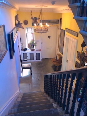 Eine Treppe führt zu einer Empore mit einem Chandelier, gerahmten Kunstwerken und einer Tür mit Wappen.