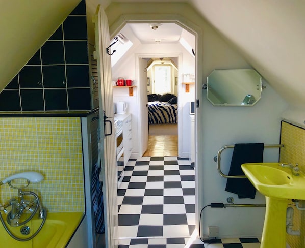 A narrow hallway leading to a bedroom with a checkered floor, a bed with a zebra-striped cover, and a small kitchenette with a kettle.