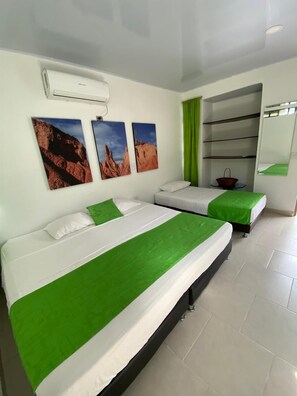 Une chambre d’hôtel avec deux lits, une couvre-lit verte et blanche, un climatiseur fixé au mur et une étagère avec un panier.