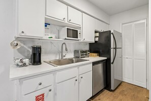 Cozinha compacta com armários brancos, pia de aço inoxidável, micro-ondas, geladeira preta e lava-louças.