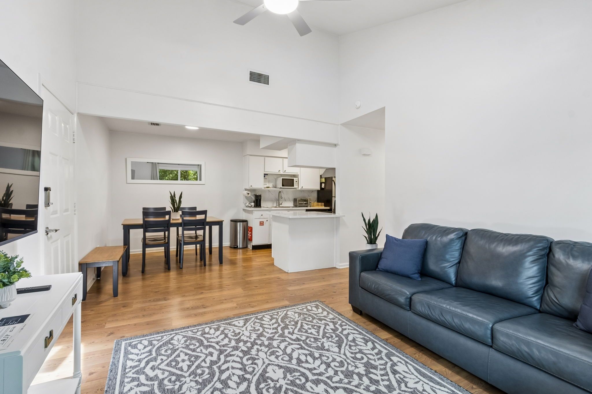 A modern living room with a dark leather sofa, a dining area with a table and chairs, and a kitchen with white cabinetry and appliances.
