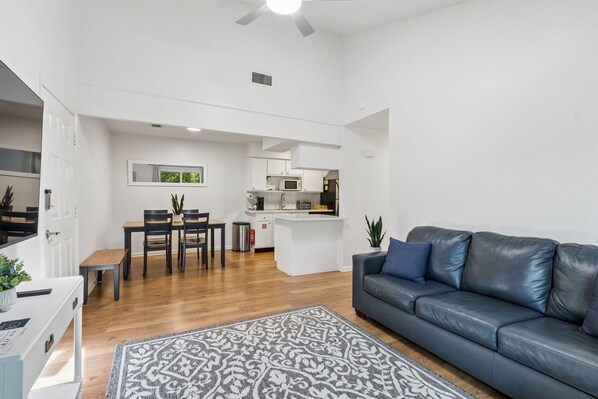 A modern living room with a dark leather sofa, a dining area with a table and chairs, and a kitchen with white cabinetry and appliances.
