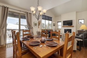 A dining area with a wooden table set for six, a chandelier, and a view of a snowy landscape through large windows. - Trailhead Lodge 734 2 Bedroom Condo by RedAwning (Winter Park)