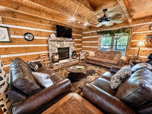 A log cabin interior with a stone fireplace, leather sofas, and a ceiling fan. - Dogwood Acre 3 Bedroom Cabin by RedAwning (Sevierville)