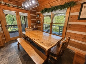 Interior de madera con una mesa de comedor y bancos, ventanas amplias con vistas a la vegetación y una lámpara de techo.