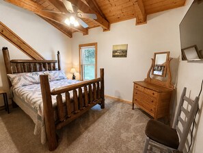 Un dormitorio con cama de madera, cómoda, silla, ventilador de techo y ventana.