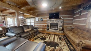 A cozy log cabin interior with a stone fireplace, leather sofas, and a television mounted above it.