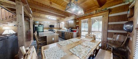 A rustic cabin interior with a wooden dining table, chairs, a kitchen area, and a fireplace.