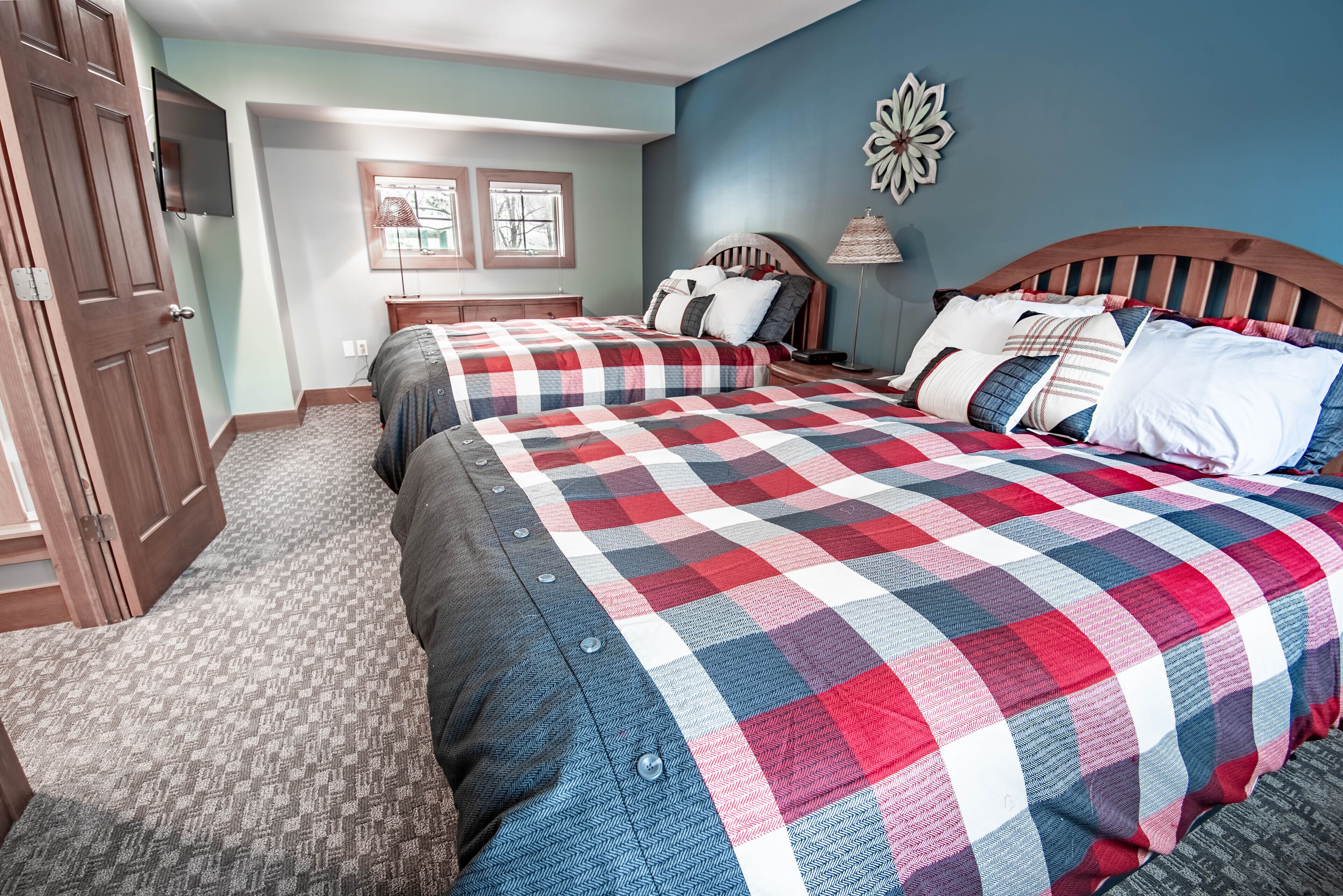 Une chambre avec deux lits, une tête de lit en bois, une table de chevet, une lampe et un téléviseur fixé au mur.