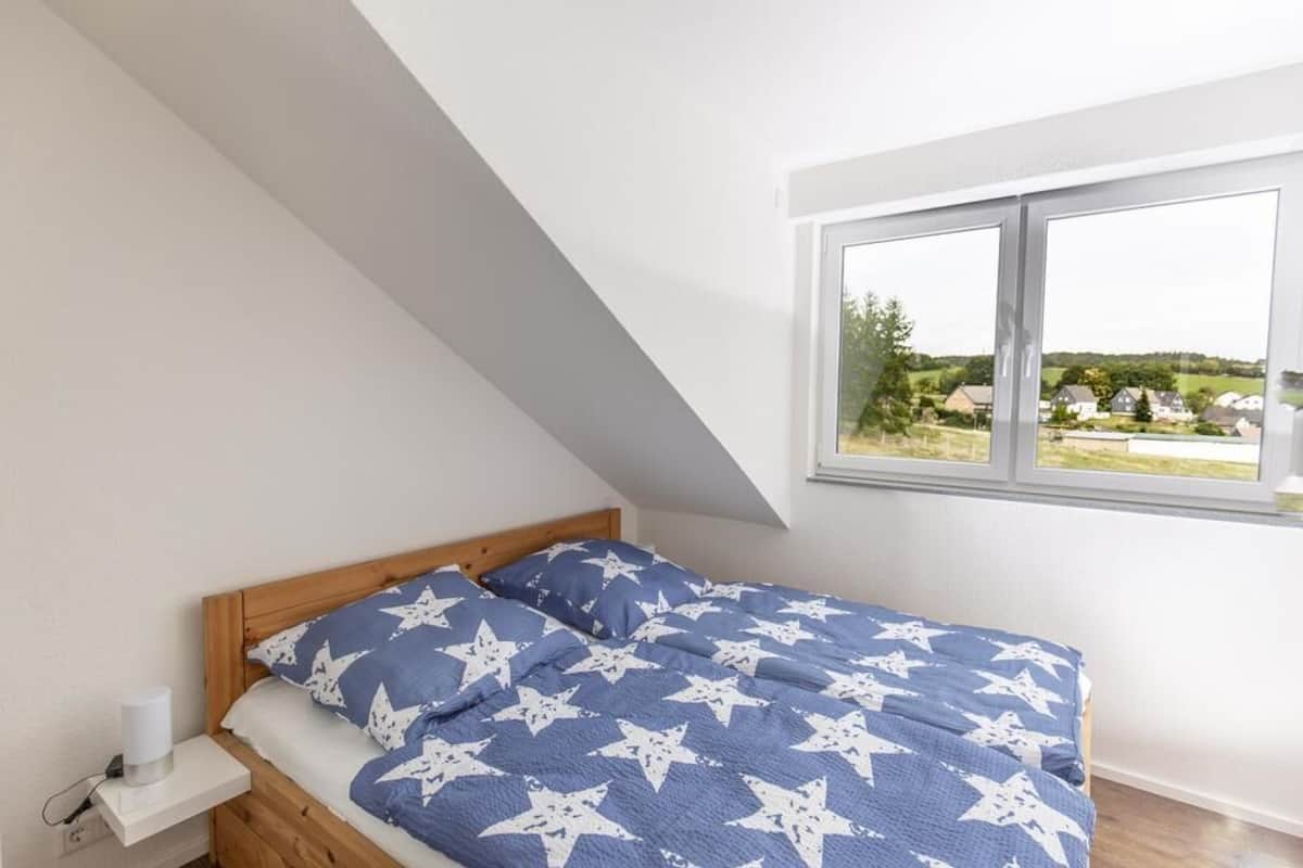 A bedroom with a bed, a nightstand, and a window with a view of houses and greenery.