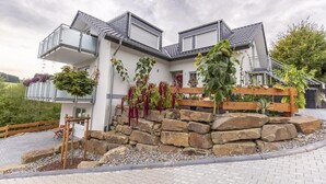 Uma casa moderna de dois andares com varanda, degraus de pedra e um jardim com diversas plantas.