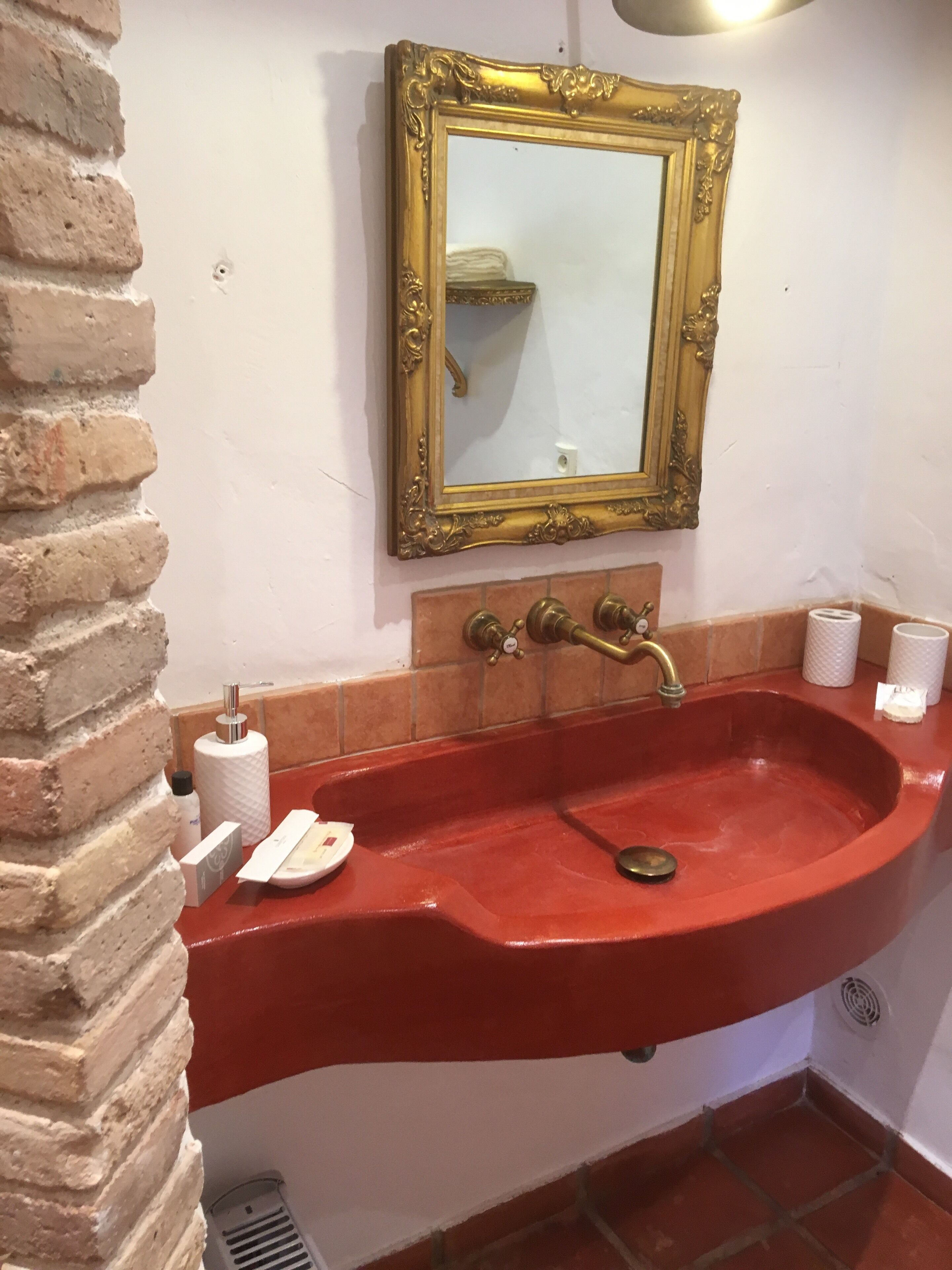 A red sink with a gold ornate mirror above it, a brick wall, and a tiled backsplash.