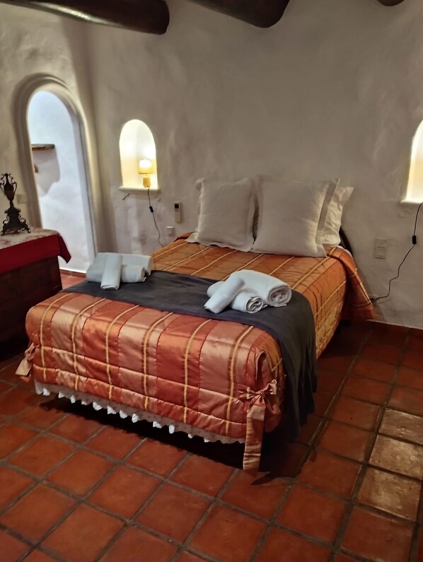 Ein Bett mit kariertem Überwurf und Kissen, ein Nachttisch mit Lampe sowie ein Fenster mit Blick ins Freie.