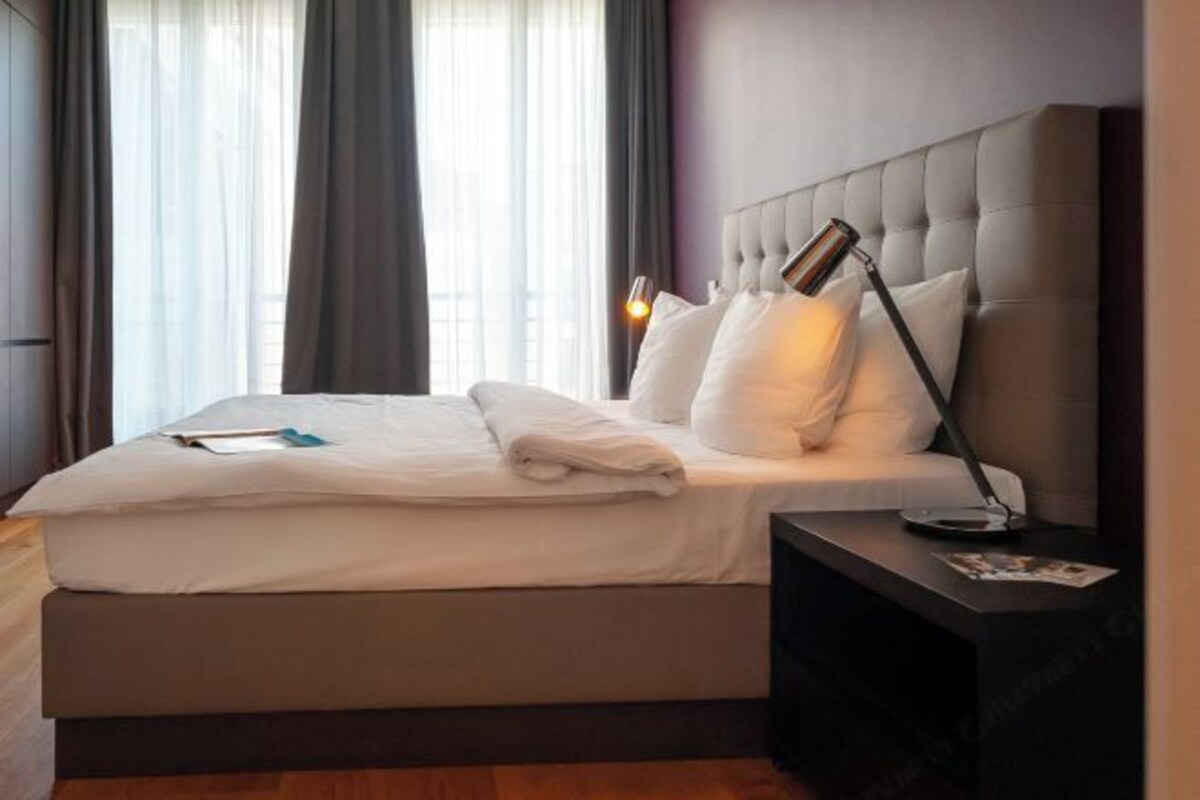 Una cama bien hecha con sábanas blancas, una lámpara de noche y una mesita de noche con una revista.