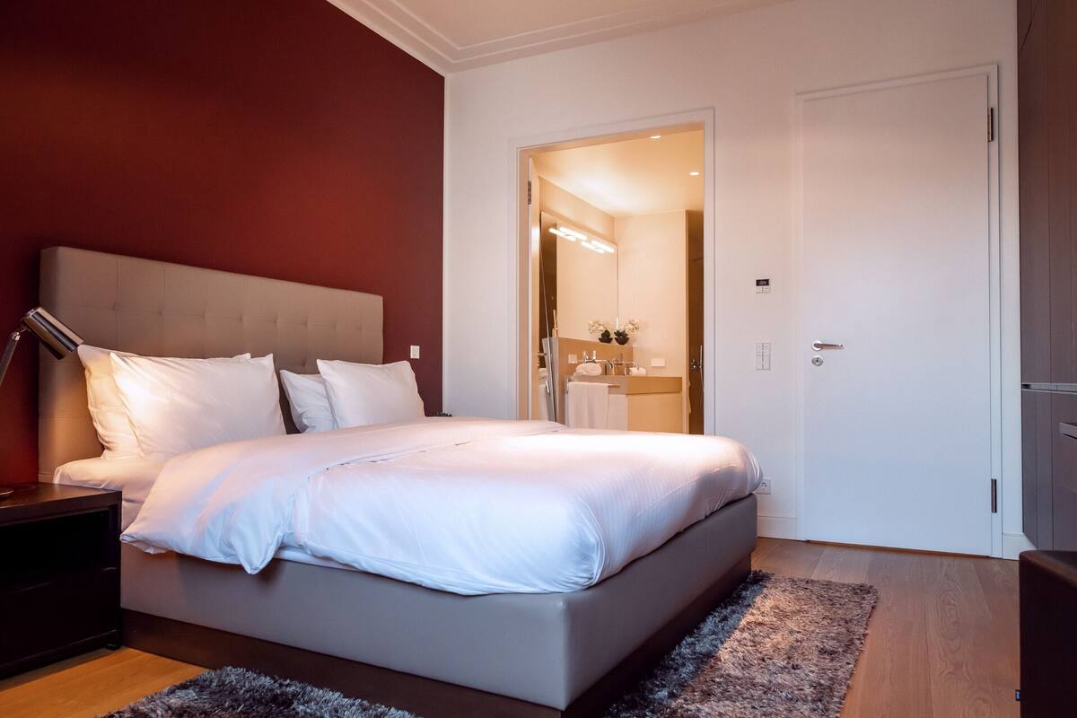 Quarto de hotel moderno com uma cama grande, uma mesa de cabeceira, uma luminária e vista para o banheiro através de uma porta aberta.