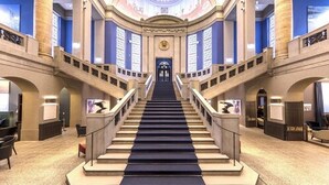 Una imponente escalera con alfombra azul que lleva a una puerta al final, flanqueada por columnas y arcos.