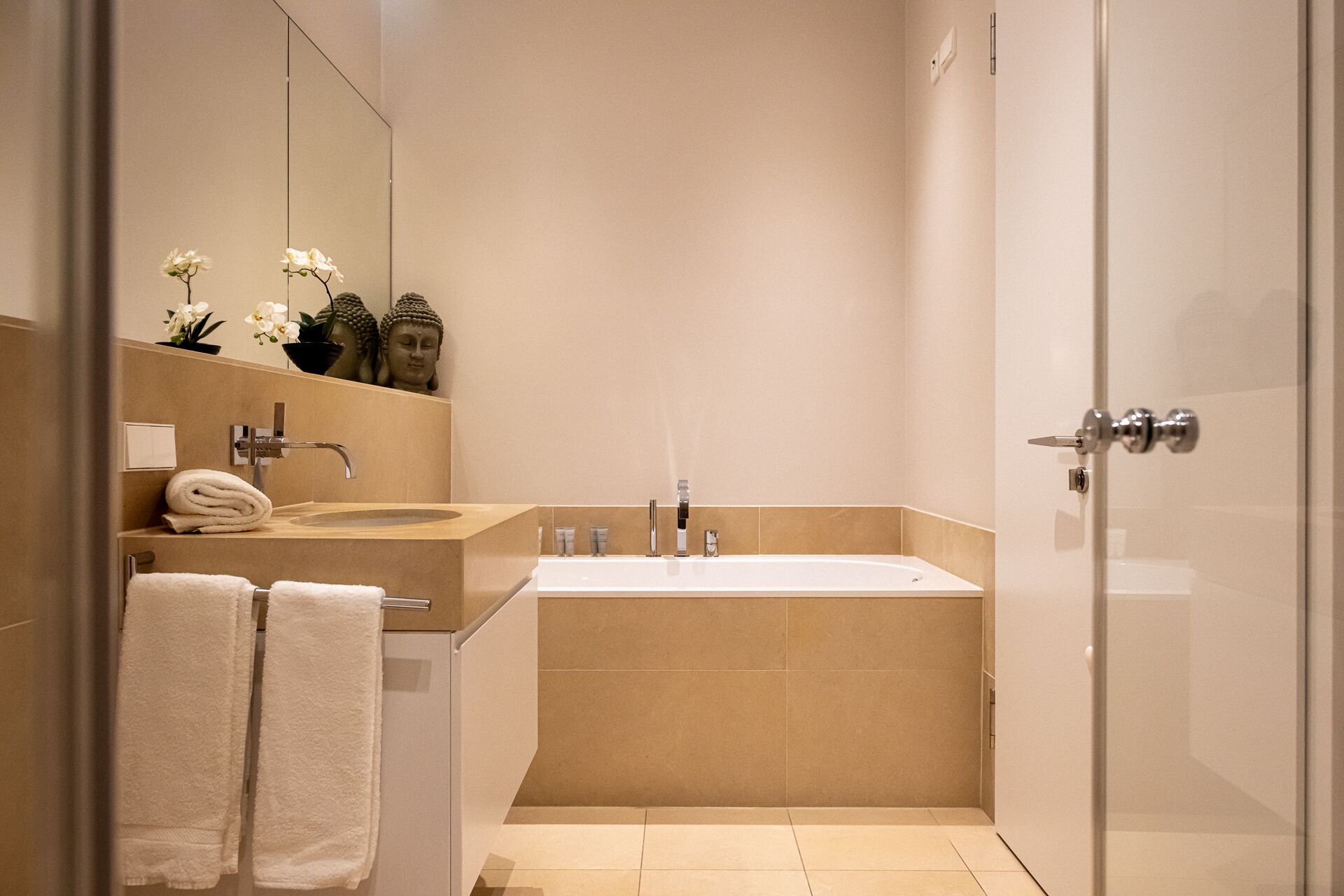 A modern bathroom with a large bathtub, a sink, and a mirror. There are towels and a Buddha statue on the counter.