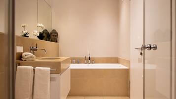 A modern bathroom with a large bathtub, a sink, and a mirror. There are towels and a Buddha statue on the counter.