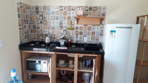 A compact kitchen with a gas stove, microwave, and a white refrigerator. A wooden shelf unit with dishes and a water dispenser is visible.