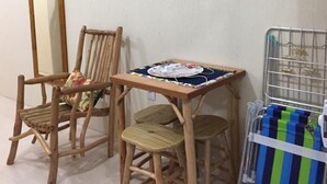 A room with a television mounted on the wall, a wooden chair with a cushion, a small table with a colorful cloth, and a metal rack with items.