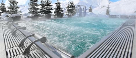 Uma nascente termal com deck de madeira e grades de metal, rodeada por montanhas cobertas de neve e árvores perenes.