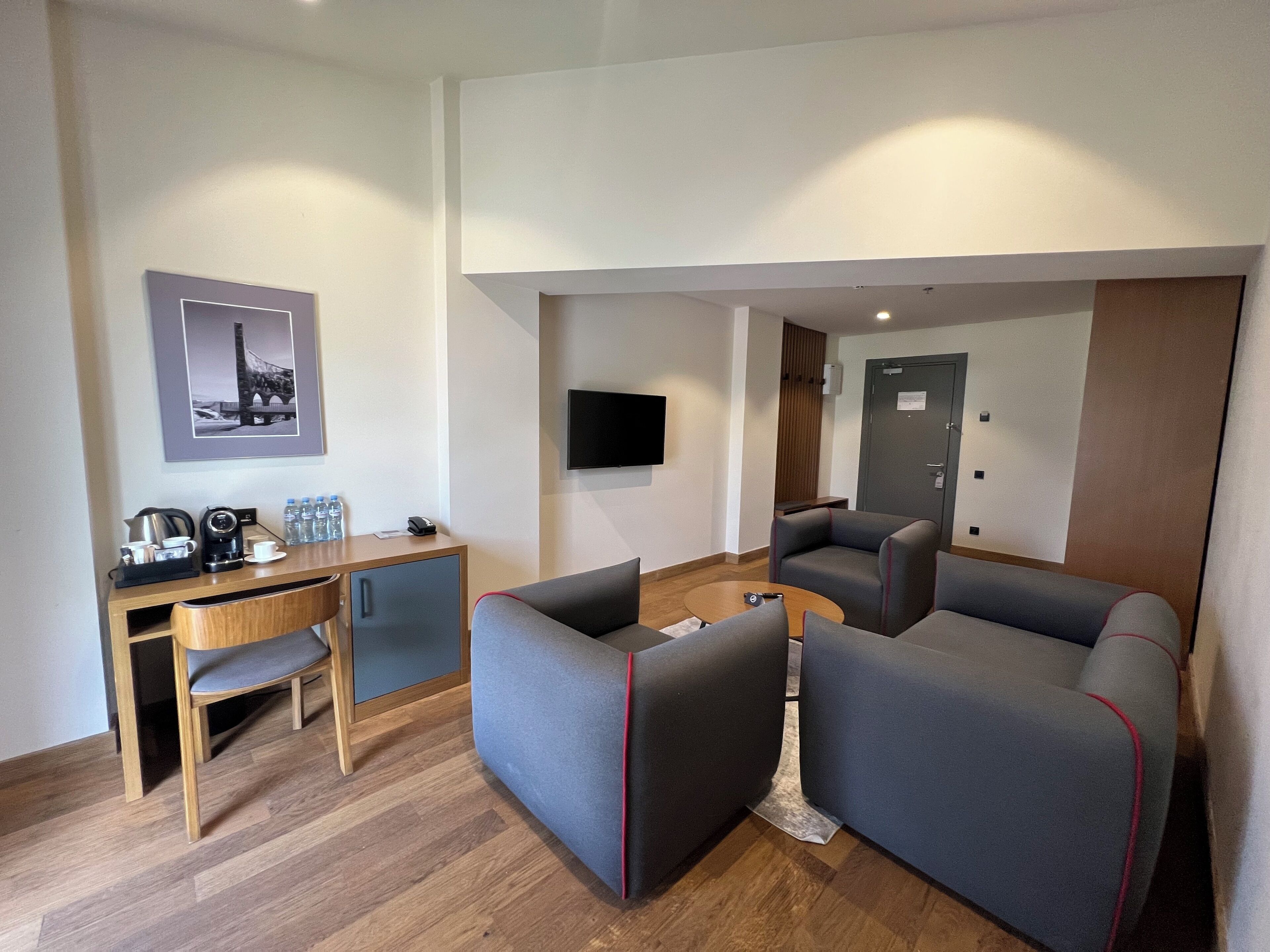 Una habitación de hotel moderna con un escritorio de madera, un sofá, un televisor montado en la pared y una puerta con cerradura.