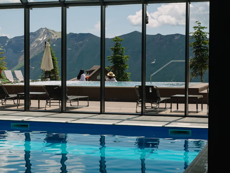 An indoor swimming pool with clear mountain views, lounge chairs, and a small building with a roof.