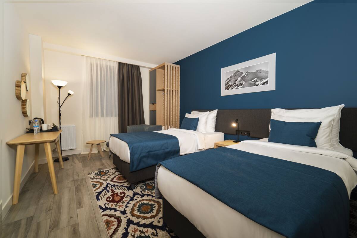 Habitación de hotel con dos camas, un escritorio, una silla, una lámpara y un gran ventanal con cortinas.