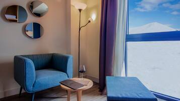 A modern room with a blue sofa, a small wooden table with a laptop, a floor lamp, and a view of a snowy landscape through a window.