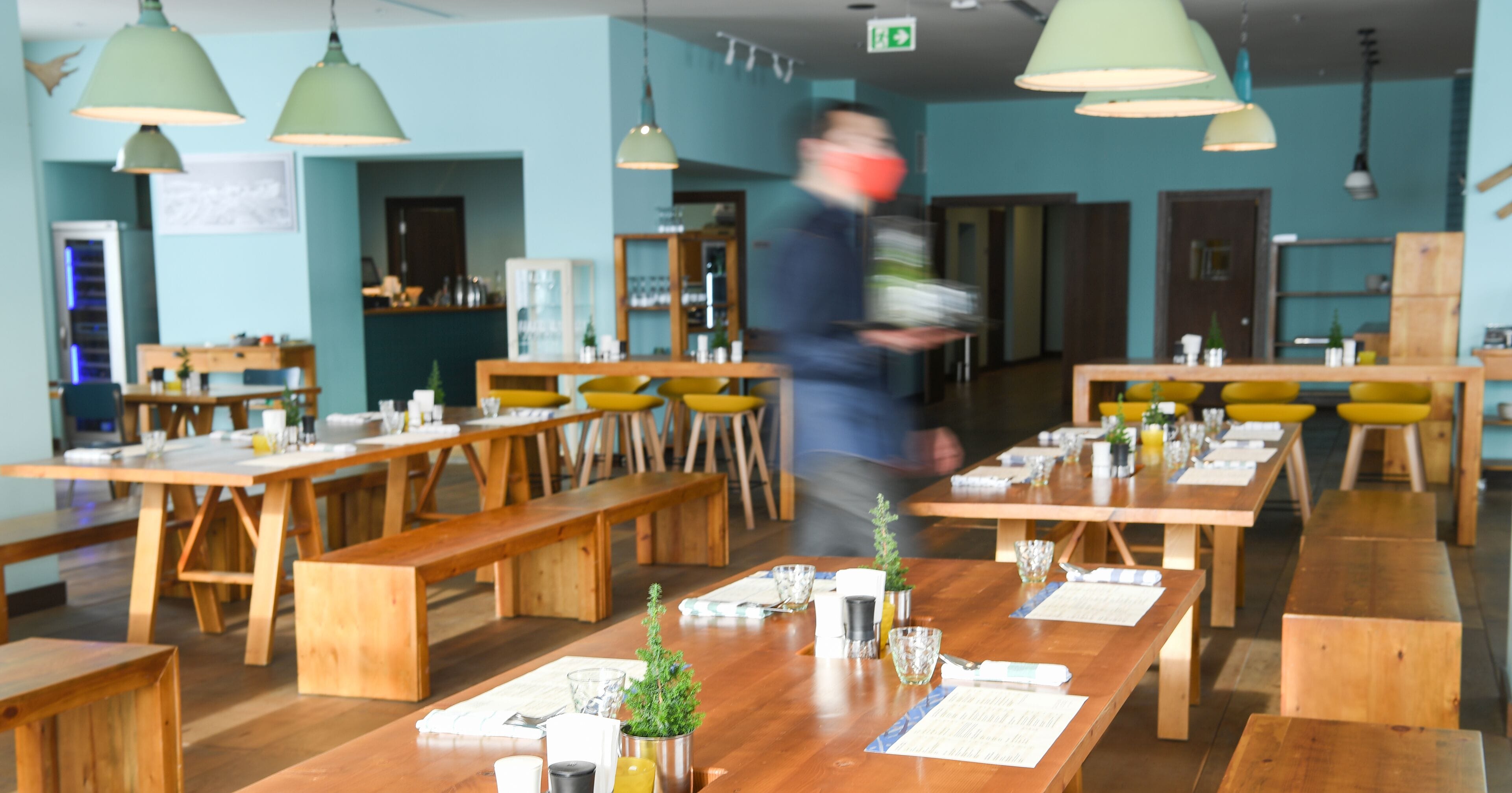 A restaurant with wooden tables and benches, green pendant lights, and a person walking through.