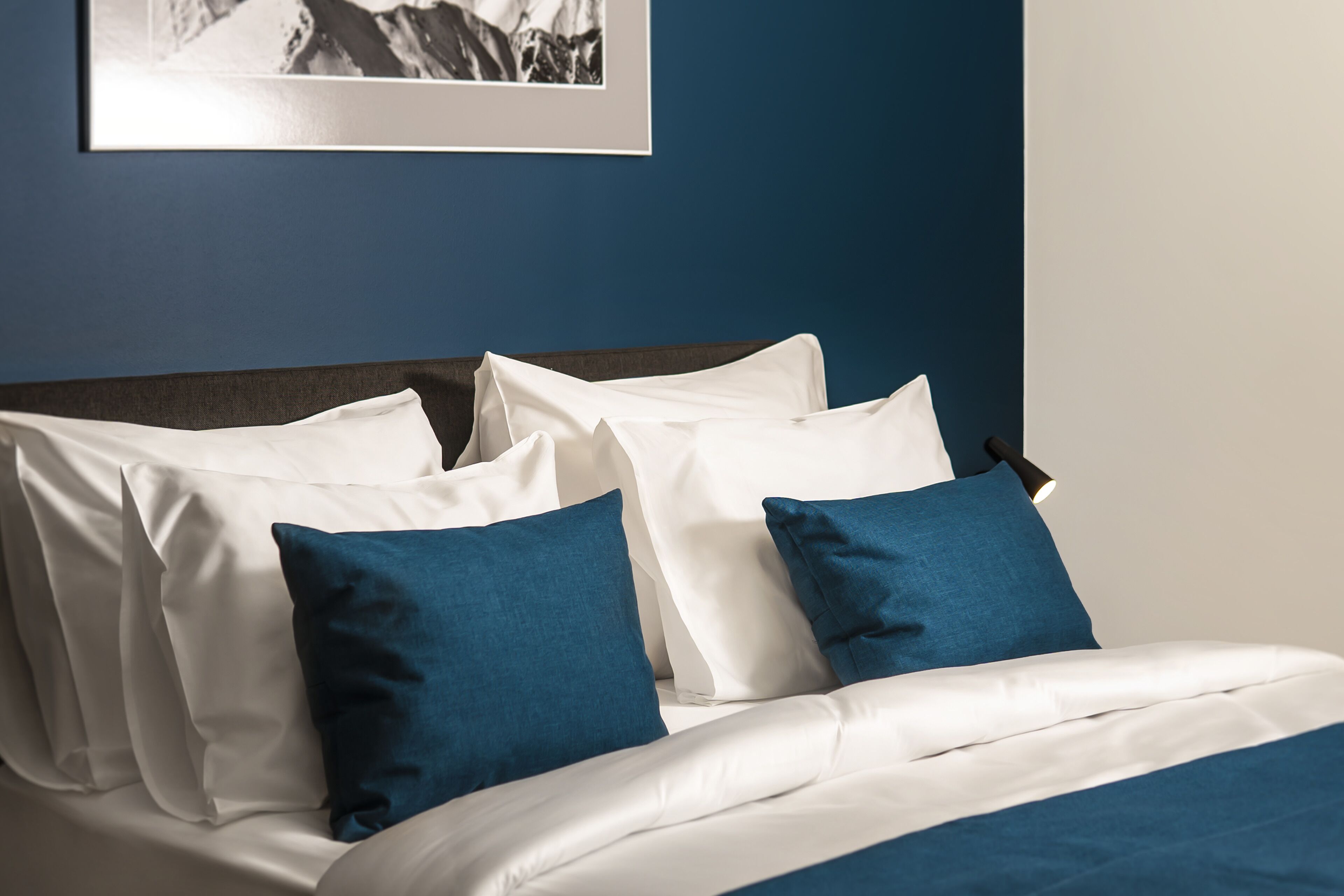 Una cama bien hecha con ropa de cama blanca y azul, un cabecero oscuro y un cuadro enmarcado en la pared.