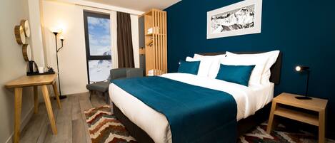 A hotel room with a bed, a desk, a chair, a nightstand, a lamp, a window with curtains, and a wall-mounted picture.