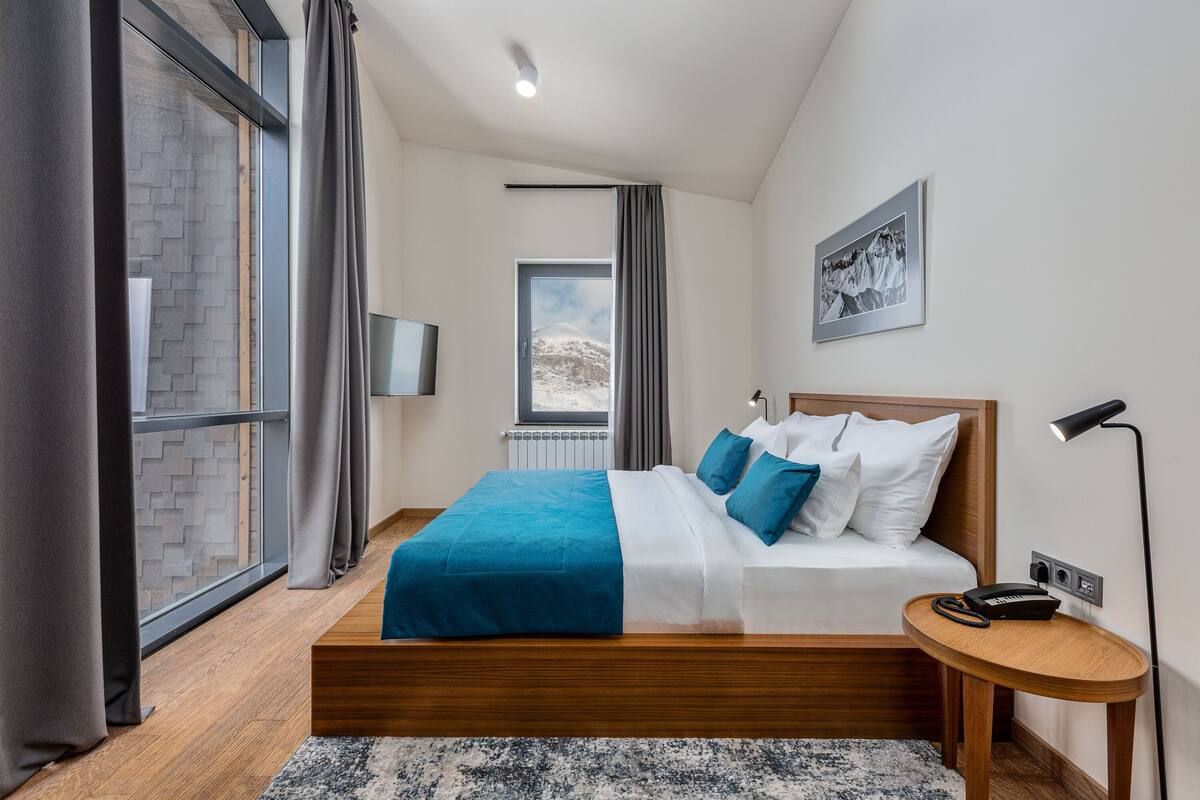 Una habitación de hotel moderna con una cama grande, una mesita de noche pequeña, una ventana con cortinas y un televisor instalado en la pared.