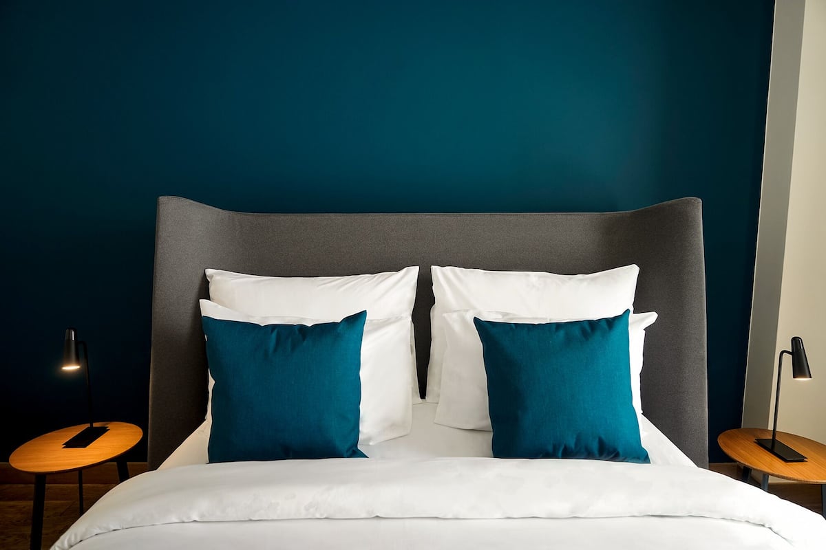 Una cama bien hecha con almohadas blancas y turquesas, un cabecero tapizado en gris y dos mesitas de noche de madera con lámparas.