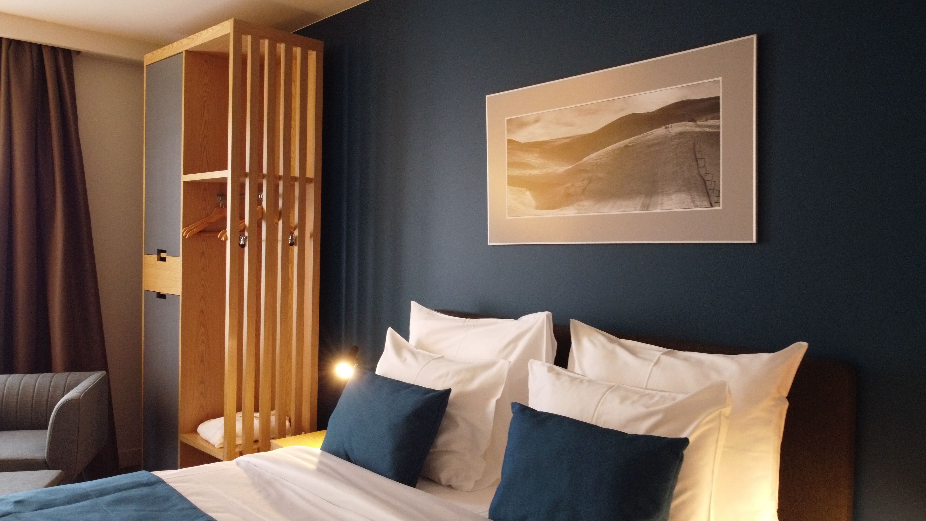 A hotel room with a bed, a chair, a wooden wardrobe, and a framed picture on the wall.