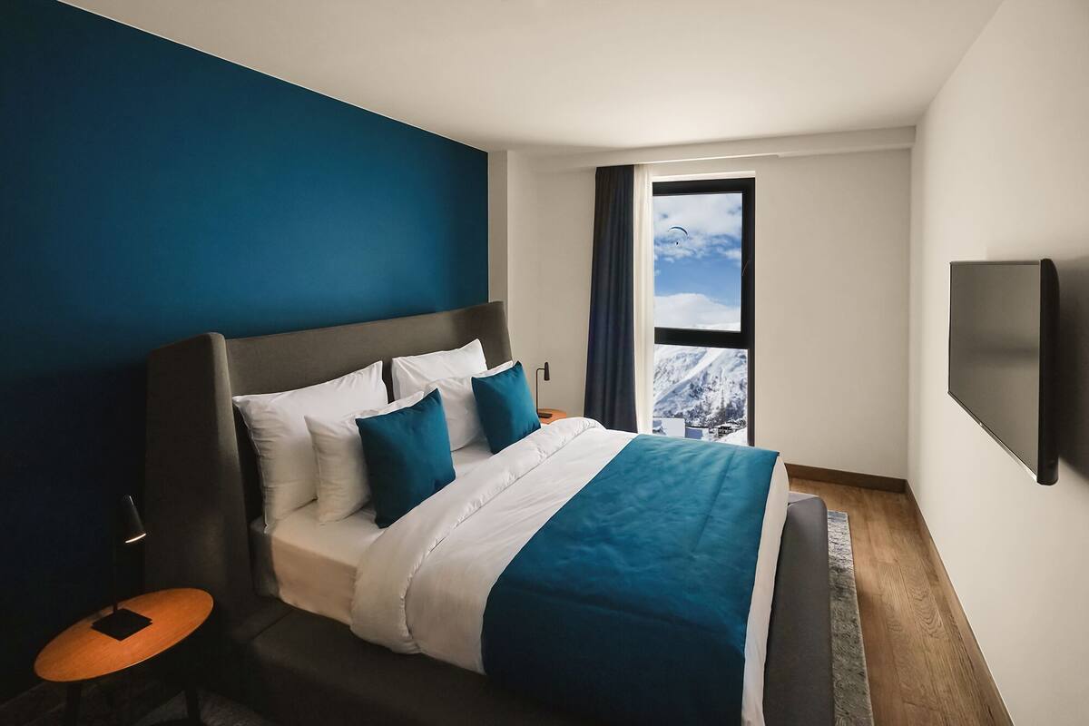 Una habitación de hotel moderna con una cama grande, vistas a montañas nevadas y un televisor montado.