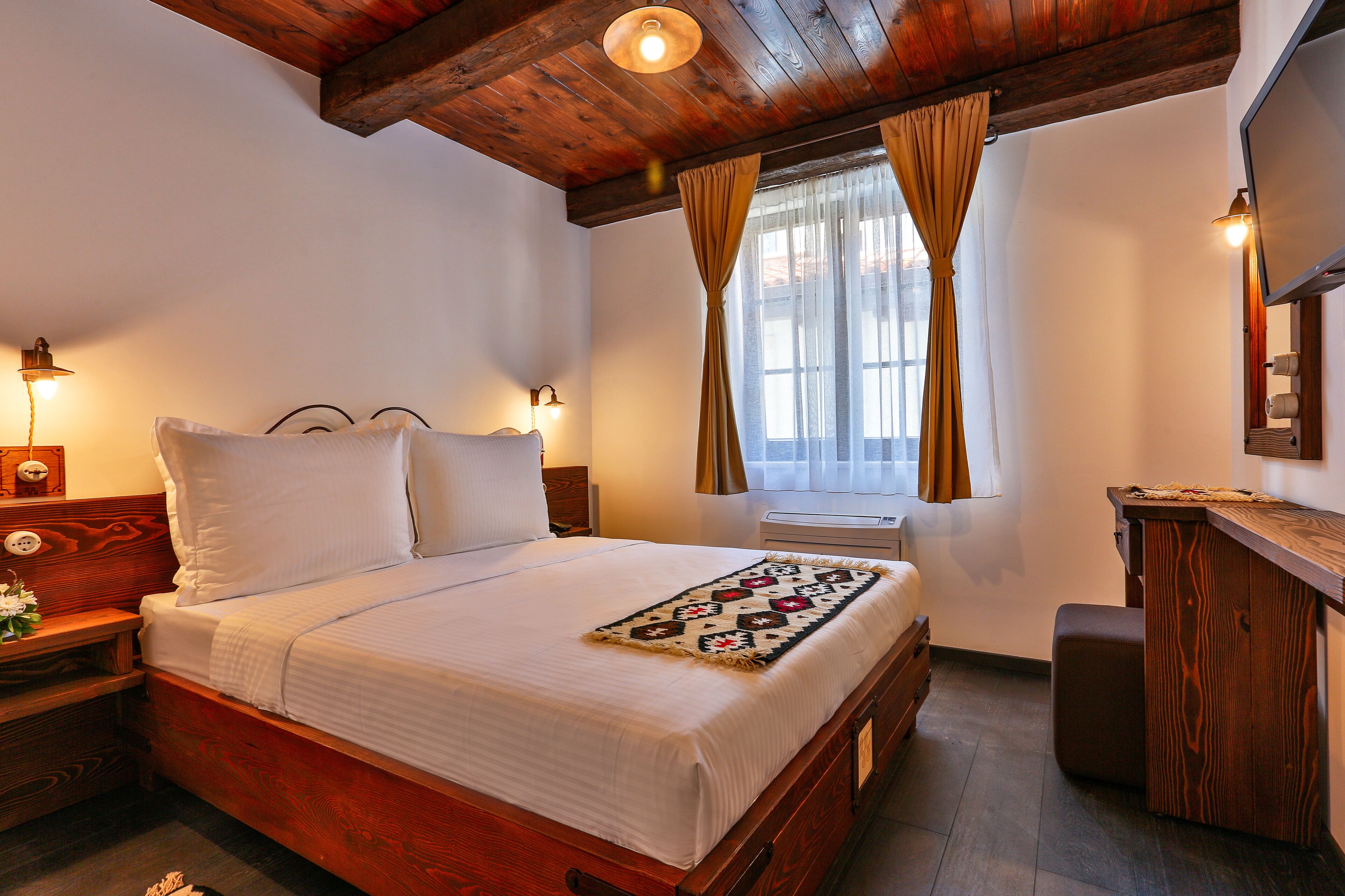 A bedroom with a wooden bed, a nightstand, a desk, and a window with curtains.