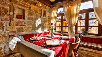 A rustic dining area with stone walls, wooden beams, a red tablecloth, and a view of the countryside through a window.