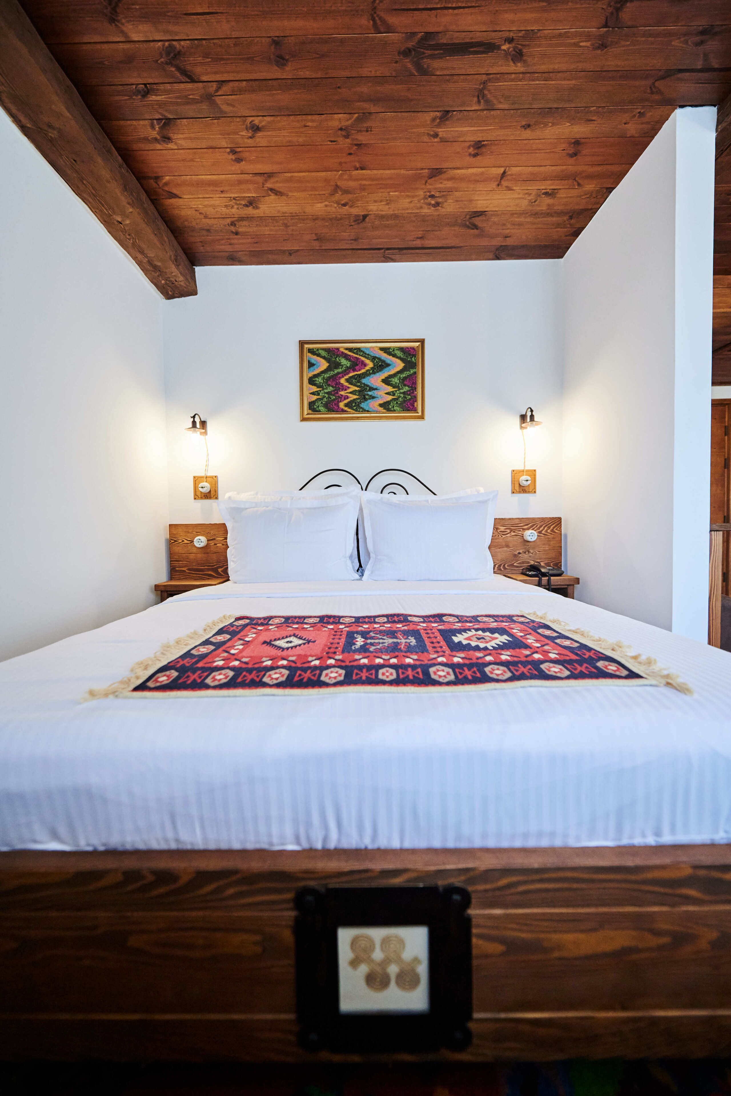 A bedroom with a wooden ceiling, a bed with white linens, two bedside tables with lamps, and a colorful wall hanging.