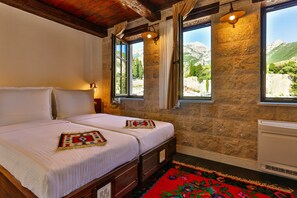 A double bed with white linens and a colorful throw blanket, a stone wall, and a window with a mountain view.