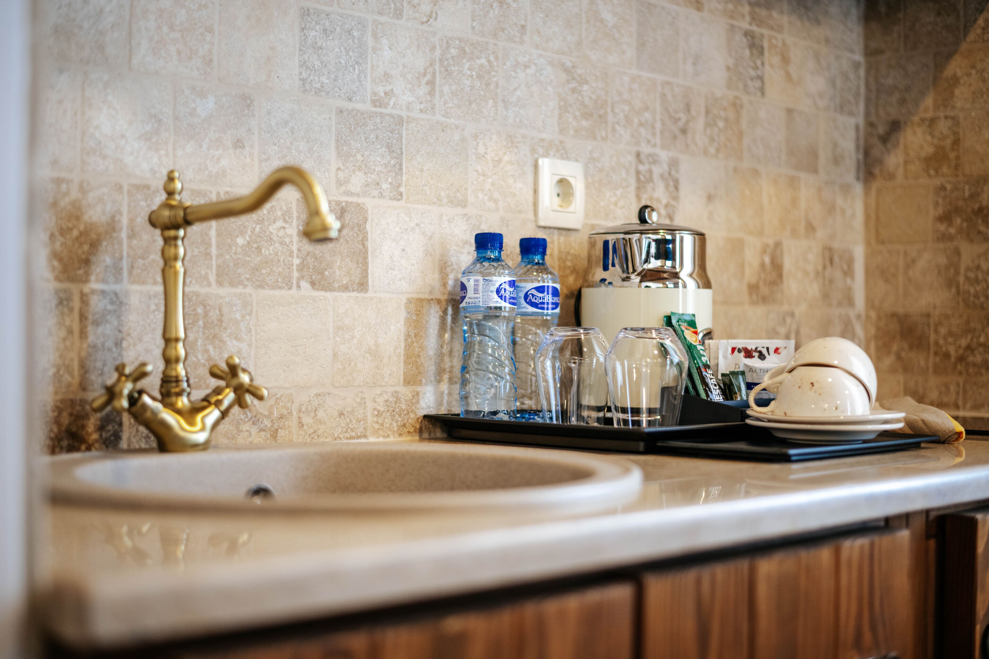 A kitchen with a vintage brass faucet, a tiled backsplash, and a countertop with water bottles, a coffee maker, and a tea kettle.