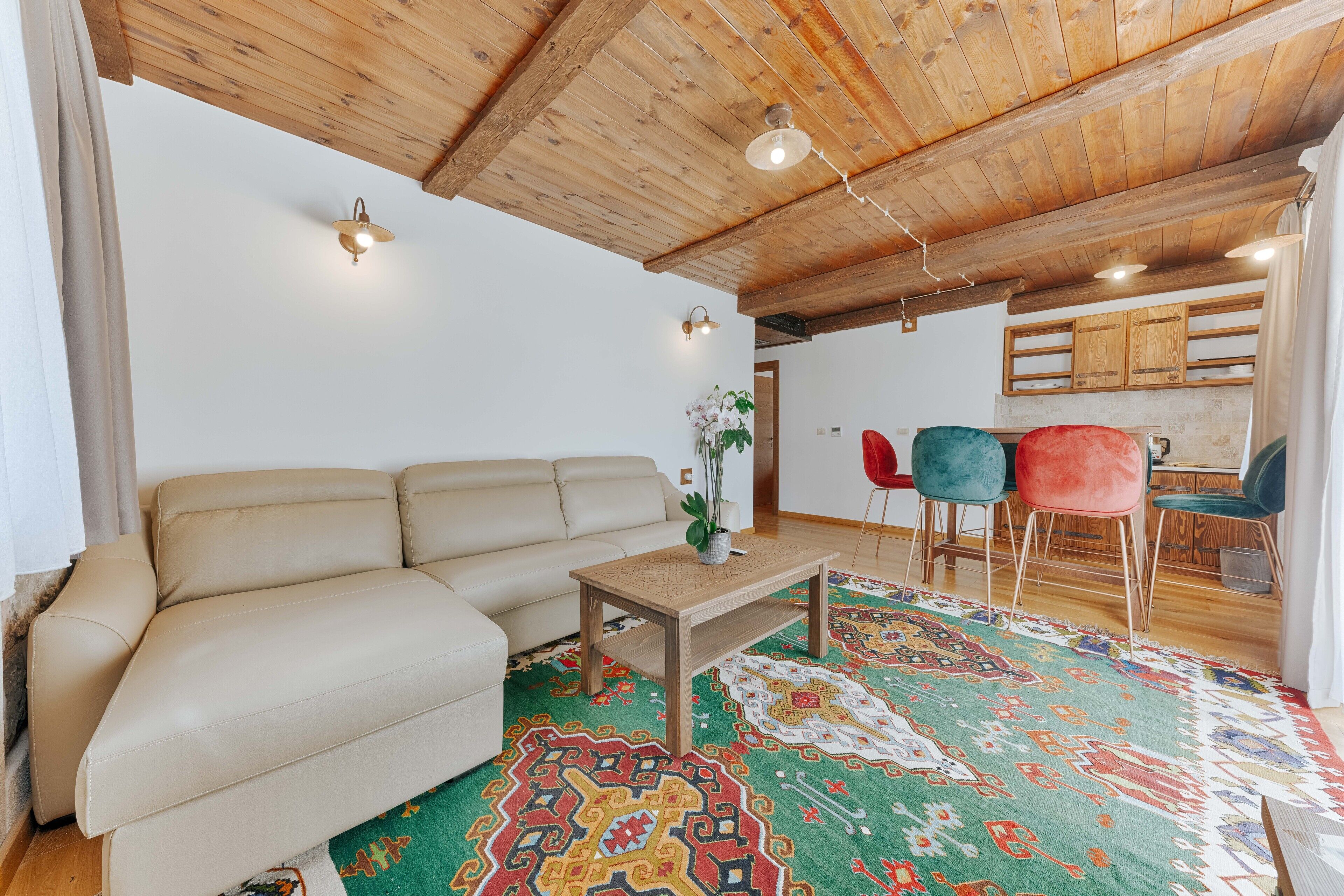 A living room with a beige sofa, a wooden coffee table, and a colorful rug. There are two chairs and a small table with a potted plant. The ceiling is wooden with exposed beams and recessed lighting.