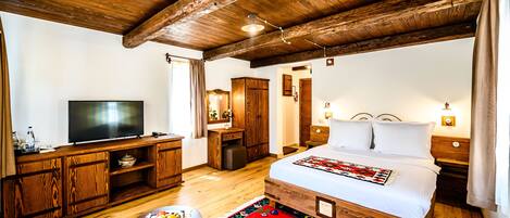 Quarto com teto de madeira, cama com roupa de cama branca, televisão sobre um móvel de madeira e uma pequena mesa com frutas.