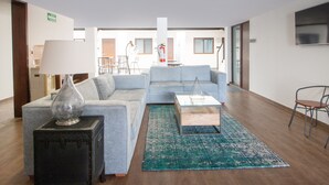 A modern living room with a grey sofa, a coffee table, a lamp, and a suitcase. There is a dining area in the background with chairs and a table. - Fraga Hotel Boutique (Irapuato)