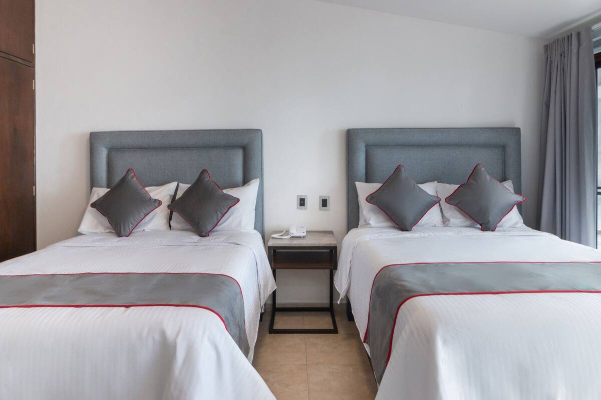 A hotel room with two beds, each with a grey headboard, white bedding, and grey pillows with red trim. There is a nightstand between the beds with a phone and a small table.