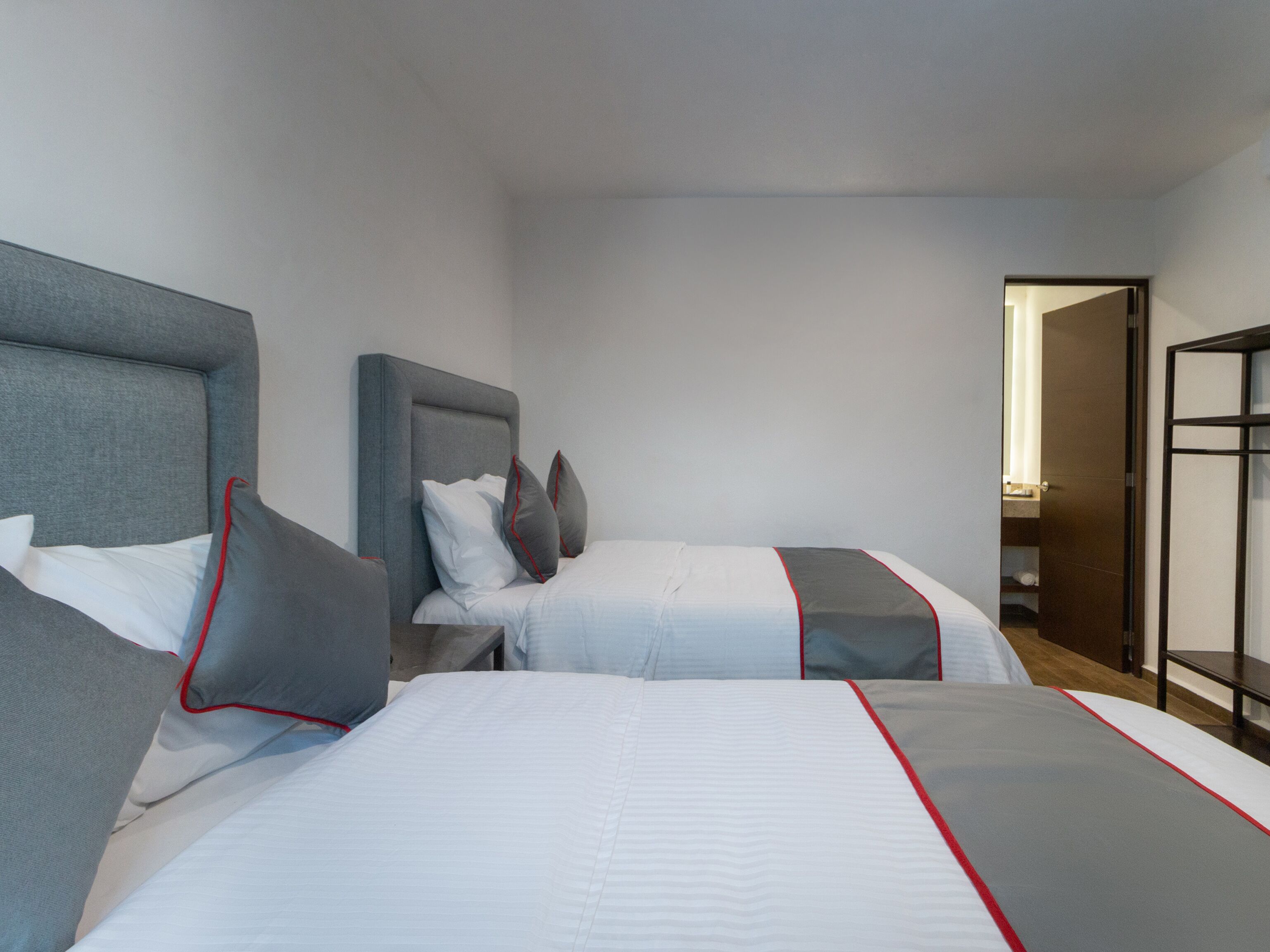 A hotel room with two beds, each with grey and white bedding and red trim.